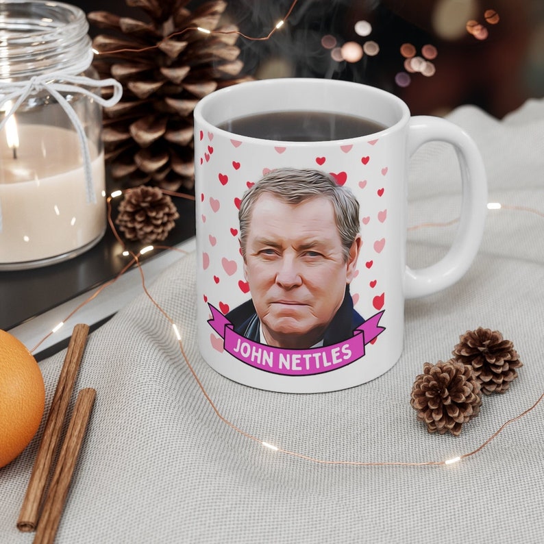 May include: White ceramic mug with a pink heart pattern and a cartoon image of John Nettles. The mug has a pink ribbon with the text "John Nettles" on it.
