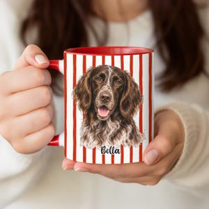 May include: A white ceramic mug with a red handle and interior, featuring a vertical red striped pattern. The center displays a detailed illustration of a brown and white spaniel dog with its mouth open, with the name Bella printed in black script.