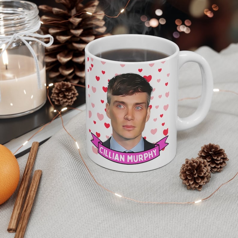 May include: White ceramic mug featuring a portrait of Cillian Murphy, surrounded by red and pink hearts. The actor's name is displayed on a pink banner. The mug is filled with a dark liquid.