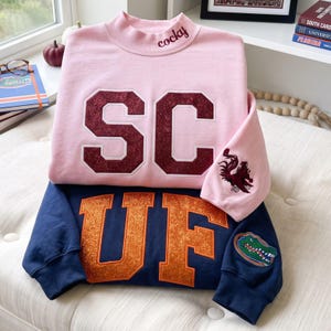 May include: A light pink mock neck sweatshirt with the word cocky embroidered on the collar and maroon glittery SC letters on the front, layered over a navy blue sweatshirt with orange glittery UF letters and a green alligator patch on the sleeve.