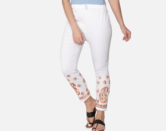 May include: A woman wearing a light blue camisole and white pants. The white pants have a colorful floral and paisley design at the ankles. The woman is wearing black sandals.