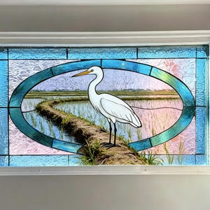 May include: Stained glass window featuring a white egret standing on a path in a rice field. The egret has a long neck and beak. The oval-shaped scene is framed by a teal and blue border, set within a white rectangular frame.