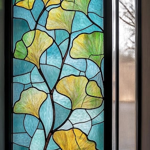May include: Stained glass window with a ginkgo leaf pattern. The leaves are yellow and green against a blue backdrop. The window is framed in black, with a view of the outside visible. The window measures approximately 100 cm x 50 cm.