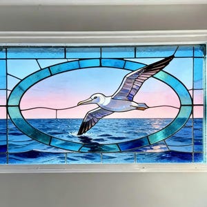 May include: Stained glass window featuring a flying seabird over a blue ocean. The bird is white and gray with black wingtips, set against a pink and blue sky. The window is framed in white.