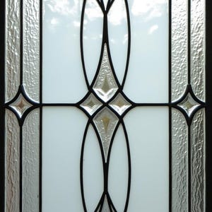 May include: A decorative glass window with a black metal frame. The design features clear textured glass panels and white opaque glass with black and silver decorative elements. The window has a symmetrical, Art Deco style.