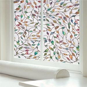 May include: A window pane with a decorative film showing a colourful leaf and vine pattern. The leaves are in shades of green, yellow, red, and brown, against a frosted background. The window is framed in white, with a white bolster pillow on the sill.