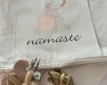 Puede incluir: Una bolsa de lona blanca con un diseño de una mujer sentada en una luna creciente con la palabra "namaste" impresa debajo. El diseño es de color cobre.
