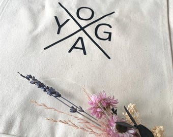 Puede incluir: Una bolsa de lona blanca con correas negras y un logotipo negro que dice "YOGA" con las letras dispuestas en forma de cruz.