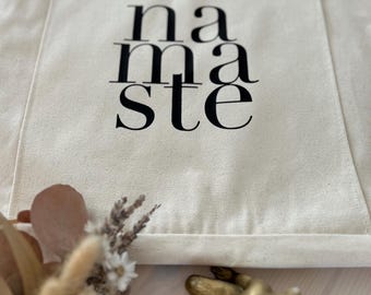 Puede incluir: Bolso tote de color crema con la palabra "namaste" impresa en negro. El bolso tiene un bolsillo en la parte delantera y dos asas. Un pequeño ramo de flores secas y una figura dorada están en primer plano.