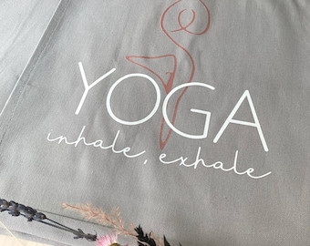 Puede incluir: Una bolsa de lona gris con un diseño de yoga blanco y el texto "yoga inhale, exhale" en blanco. El diseño de yoga es una persona estilizada en una postura de yoga.