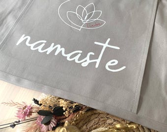 Puede incluir: Un bolso tote gris con la palabra "namaste" en escritura blanca. El bolso presenta un bolsillo con un diseño de flor de loto blanca y la palabra "Yoga" encima. Una etiqueta marrón con el logotipo "Earth Aware" está adjunta.