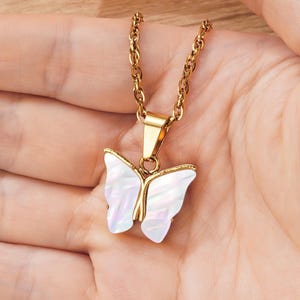 May include: A gold-colored necklace with a butterfly pendant. The pendant features a white, iridescent butterfly design with gold-colored accents. The necklace is held in a person's hand.