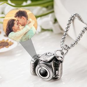 May include: A silver-toned metal camera pendant necklace featuring a detailed miniature camera charm with a central lens. A circular projection insert displays a photograph of a couple embracing. The pendant hangs from a silver-toned box chain.