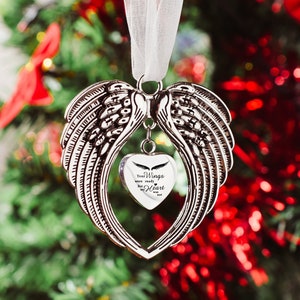 May include: Silver angel wing ornament with a heart charm that reads "Your wings were ready but my heart was not". The ornament is hanging on a white ribbon against a blurred background of a Christmas tree.