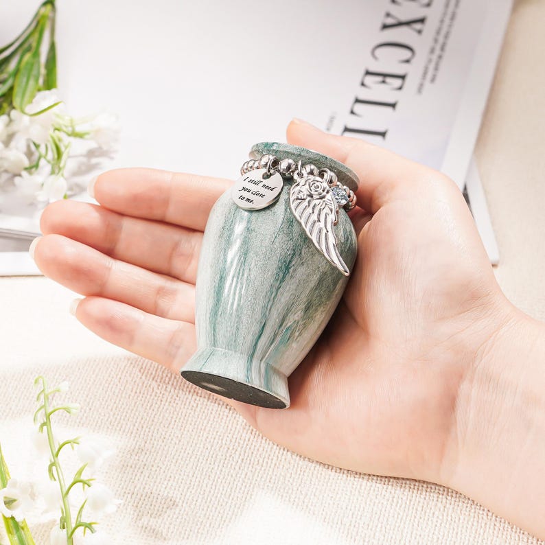 May include: A small, green and white marbled keepsake urn featuring a silver-toned charm bracelet around its neck. The bracelet includes a round metal tag engraved with the text I still need you close to me and a detailed silver angel wing charm.