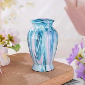 May include: A small, urn-shaped vessel featuring a glossy finish with a swirling marble pattern in shades of light blue, dark blue, and soft pink. The container has a rounded body, a narrow neck, and a flared base, topped with a matching lid.
