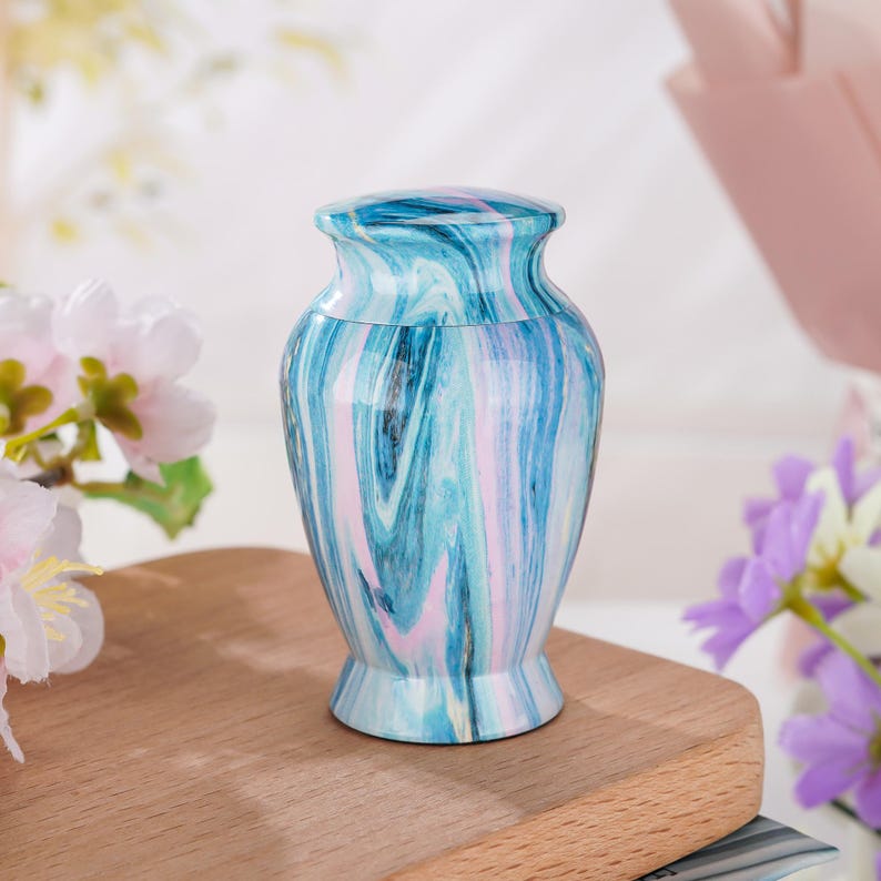 May include: A small, urn-shaped vessel featuring a glossy finish with a swirling marble pattern in shades of light blue, dark blue, and soft pink. The container has a rounded body, a narrow neck, and a flared base, topped with a matching lid.