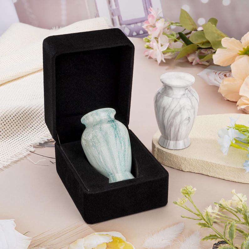 May include: A small, light green marble-patterned keepsake urn rests inside a black velvet-lined presentation box. Beside it stands a second, white and gray marble-patterned keepsake urn with a matching lid, both featuring a classic vase shape.