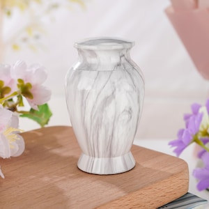 May include: A small, urn-shaped vessel featuring a smooth, polished finish with a white and grey marbled pattern. The container has a rounded body, a flared base, and a matching lid that sits flush with the top opening of the vessel.