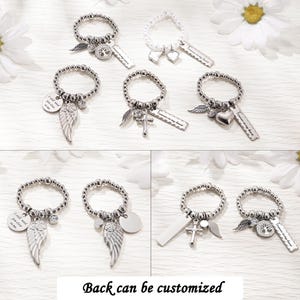 May include: A collection of beaded rings featuring silver-toned charms including angel wings, hearts, crosses, and tree of life motifs. Some rings include rectangular tags with the engraved text "Forever in my heart" or circular tags with "I still need you close to me."