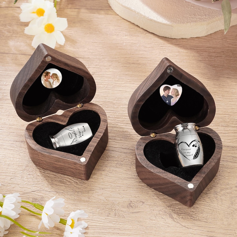 May include: Two heart-shaped wooden boxes with a small silver urn inside. The boxes are open, revealing a black interior and a small photo. One urn has the text "Loved Always" and the other has "Forever in my heart".