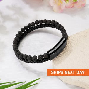 May include: Black lava stone bracelet with a braided leather band and a magnetic clasp. The bracelet features a double-strand design with small, round lava beads. The clasp is a sleek, rectangular shape.