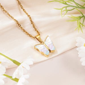 May include: A gold-toned chain necklace featuring a butterfly-shaped pendant. The pendant has a metallic gold border and wings filled with an iridescent, pearlescent material that displays subtle shifts of white, blue, and pink light reflections.