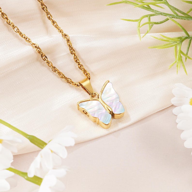 May include: A gold-toned chain necklace featuring a butterfly-shaped pendant. The pendant has a metallic gold border and wings filled with an iridescent, pearlescent material that displays subtle shifts of white, blue, and pink light reflections.