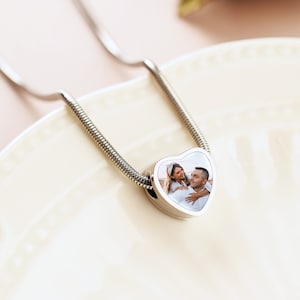 May include: A silver chain necklace with a heart-shaped pendant featuring a photo of a couple. The pendant is silver with a white background and a clear glass insert.