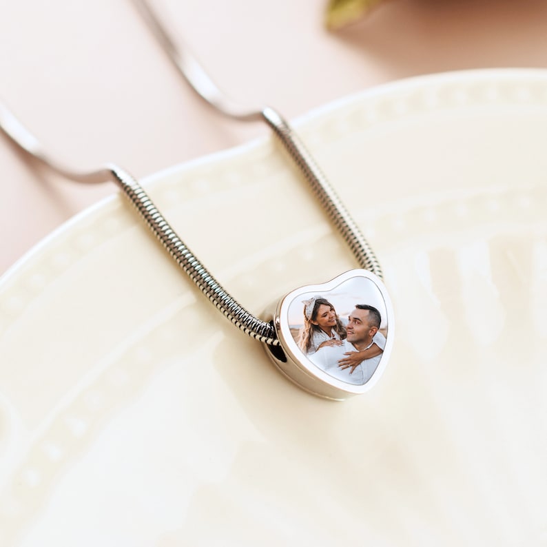 May include: A silver chain necklace with a heart-shaped pendant featuring a photo of a couple. The pendant is silver with a white background and a clear glass insert.