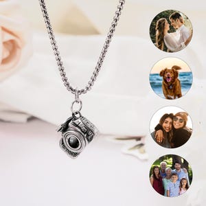 May include: A silver-toned metal camera pendant hangs from a matching box chain necklace. The camera charm features detailed engravings of lens and shutter components. To the right, four circular insets display various personal photographs.
