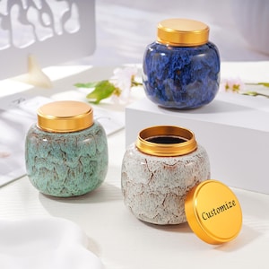 May include: Three ceramic jars with gold-colored lids. One jar is blue, one is green, and one is white with a cracked pattern. The lid of the white jar is off, revealing the text "Customize".