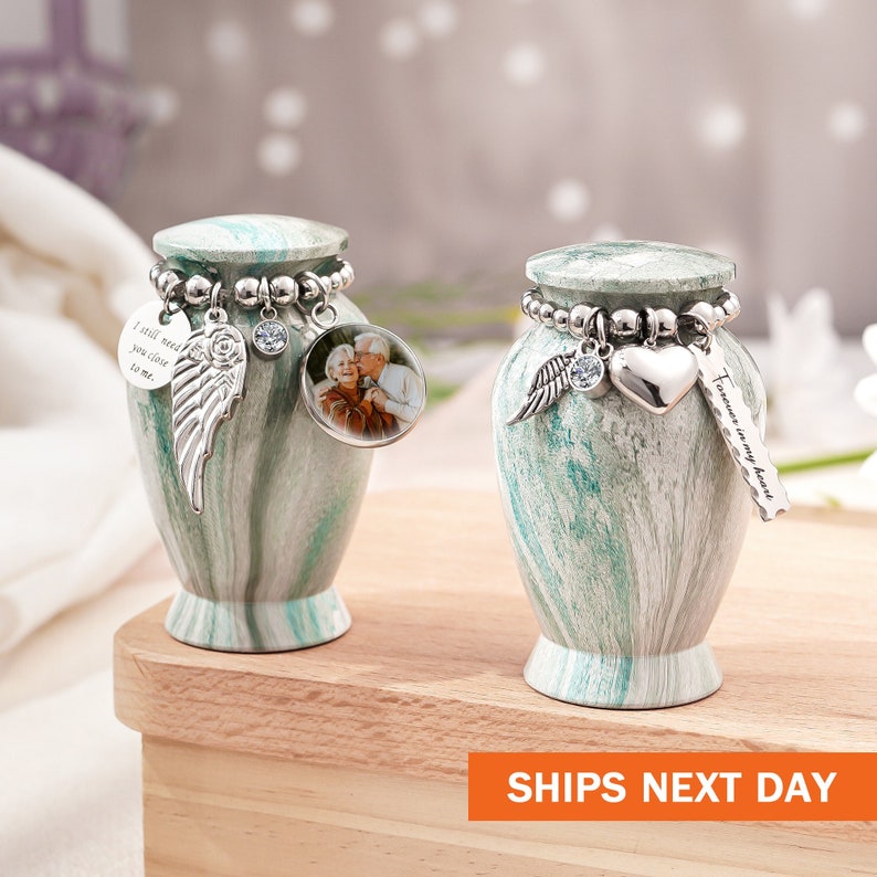 May include: Two small, green and white marbled keepsake urns with silver-toned lids. Each urn features a beaded charm bracelet around the neck. One urn has a photo charm of a couple, a wing charm, and a round disc with the text I still need you close to me. The other has a wing charm, a heart charm, and a rectangular tag with the text Forever in my heart.