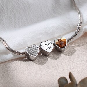 May include: A silver charm bracelet with three heart-shaped charms. The charm on the left is covered in clear rhinestones. The middle charm has the words "Forever Love" engraved on it. The charm on the right has a photo of a couple inside.