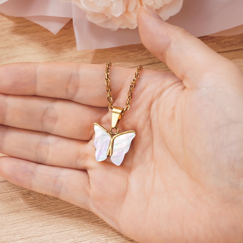 May include: A gold-colored necklace with a butterfly pendant. The pendant features a white, iridescent butterfly design with gold-colored accents. The necklace is held in a person's hand.
