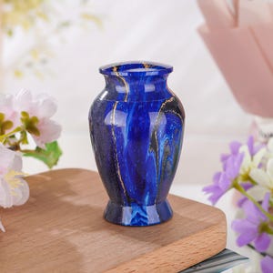May include: A small, cylindrical keepsake urn featuring a deep blue finish with swirling black patterns and thin, metallic gold-colored veins. The vessel has a narrow neck, a flared rim, and a matching lid, presenting a smooth, polished surface.