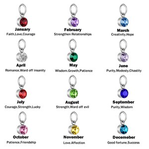 May include: A collection of twelve silver-toned metal charms, each featuring a circular gemstone in a unique color representing a month. The text below each charm lists the month and associated traits like Faith, Love, Courage, and Good fortune.