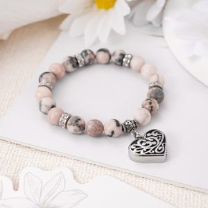 May include: A pink and white beaded bracelet with a silver heart charm. The heart charm has a filigree design.