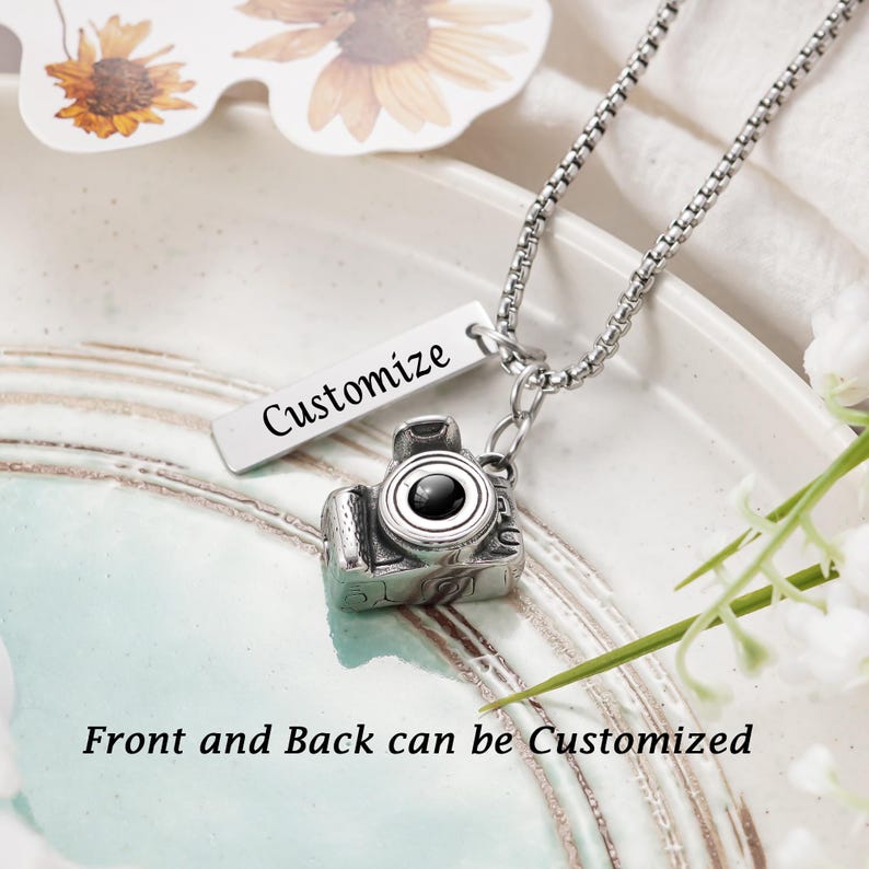 May include: A silver-toned metal necklace featuring a miniature camera pendant and a rectangular bar charm engraved with the word Customize. The text Front and Back can be Customized is visible. The chain is a box link style made of silver metal.