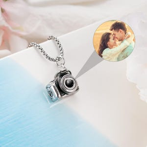May include: A silver-toned metal camera pendant necklace featuring a circular projection lens. A circular inset displays a color photograph of a couple embracing outdoors. The pendant hangs from a silver-toned box chain with a metallic finish.