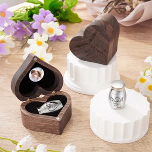May include: A collection of memorial keepsakes, including heart-shaped wooden boxes and a small silver urn. The wooden boxes feature engraved text and a photo. The urn has angel wings and text. The items are displayed on white pedestals.