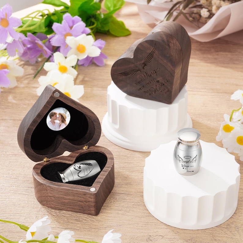 May include: A collection of memorial keepsakes, including heart-shaped wooden boxes and a small silver urn. The wooden boxes feature engraved text and a photo. The urn has angel wings and text. The items are displayed on white pedestals.
