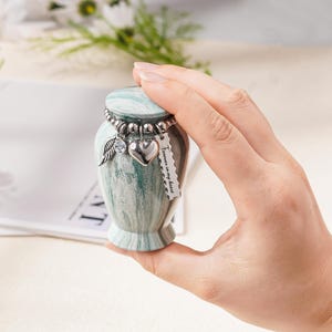 May include: A small, marble-patterned green and white keepsake urn with a removable lid. A silver-toned beaded chain encircles the neck, featuring three charms: a crystal-accented wing, a smooth heart, and a rectangular tag reading Forever in my heart.