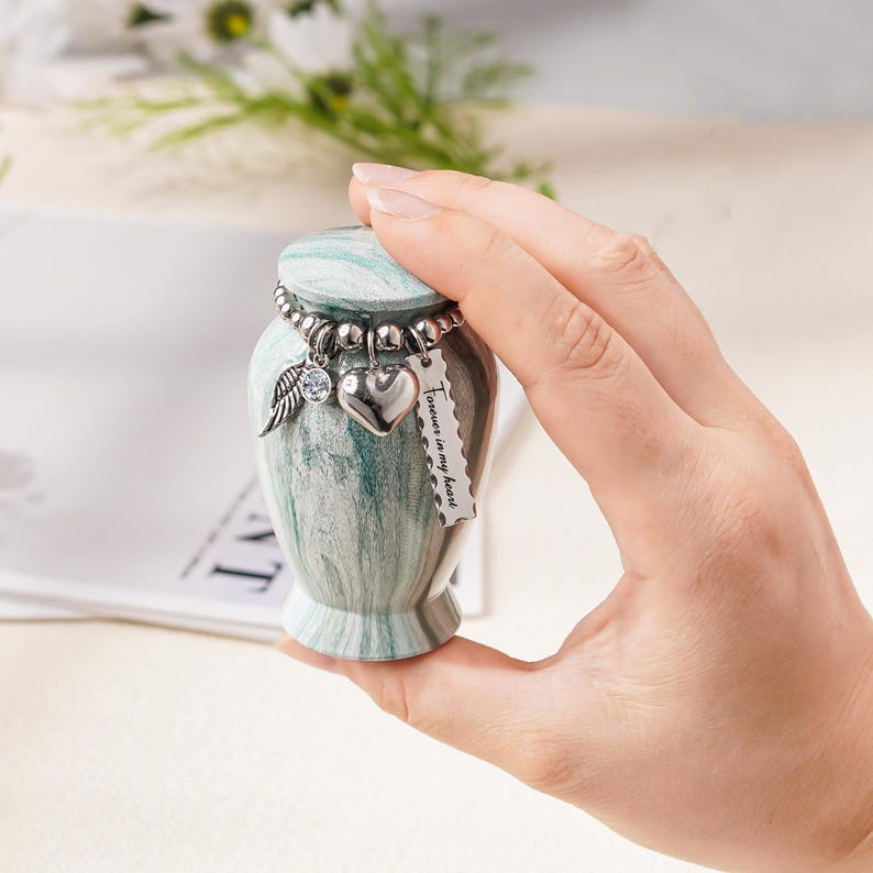 May include: A small, marble-patterned green and white keepsake urn with a removable lid. A silver-toned beaded chain encircles the neck, featuring three charms: a crystal-accented wing, a smooth heart, and a rectangular tag reading Forever in my heart.