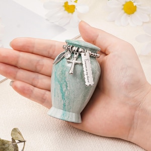 May include: A small, teal and white marbled urn featuring a silver-toned beaded chain around the neck. The chain holds three charms: a silver wing, a small cross, and a rectangular silver tag engraved with the text Forever in my heart.