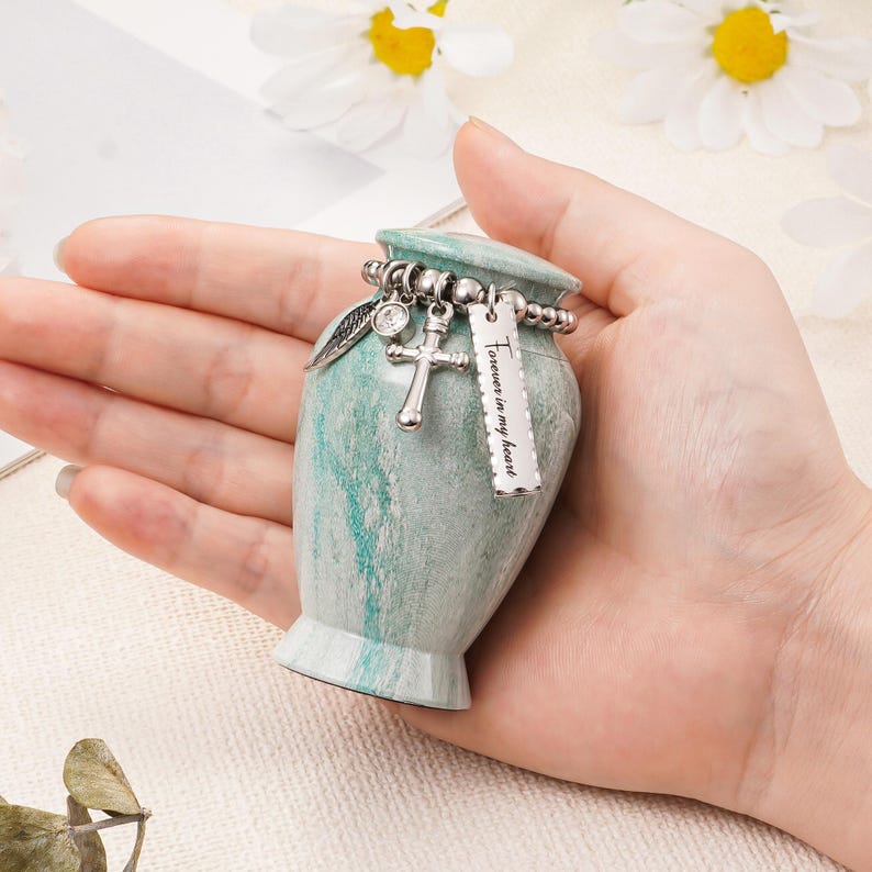 May include: A small, teal and white marbled urn featuring a silver-toned beaded chain around the neck. The chain holds three charms: a silver wing, a small cross, and a rectangular silver tag engraved with the text Forever in my heart.