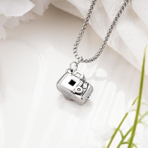 May include: A silver-toned metal necklace featuring a small, detailed camera pendant. The pendant is suspended from a sturdy box chain. The camera charm displays a viewfinder, lens, and shutter button, rendered in a polished, reflective metallic finish.