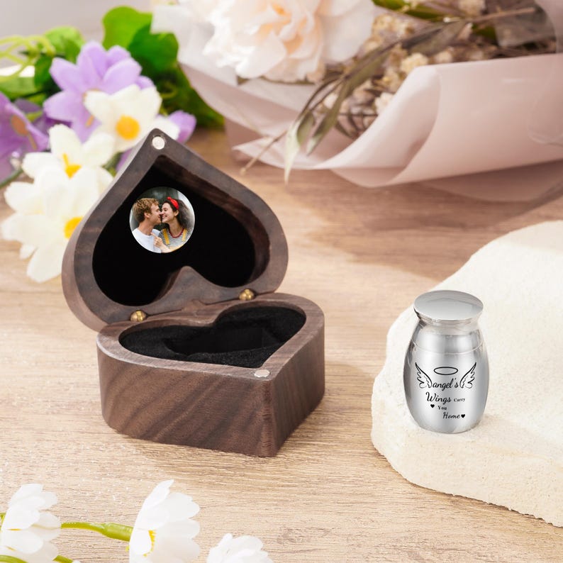 May include: A heart-shaped wooden box with a small photo inside, next to a small silver urn. The urn has the words "Angel's Wings come Home" and angel wings printed on it. The box is open, revealing a black interior.