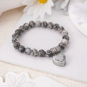 May include: A gray and white beaded bracelet with a silver heart charm that reads "Always with me".