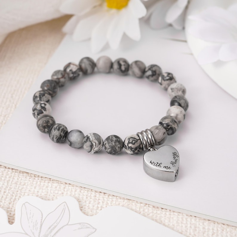 May include: A gray and white beaded bracelet with a silver heart charm that reads "Always with me".